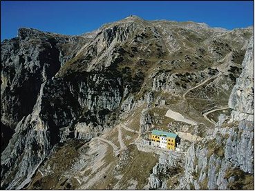 Frotte allegre di turisti sbucano dalla 52^ galleria di questa celeberrima strada, costruita in soli undici mesi, uno dei tanti assurdi miracoli prodotti dalle guerre sulle nostre montagne.Sbucano allegri e sudati, qualcuno un po stravolto, con cani e bambini, e riempiono di vita questa bellissima montagna, cos aperta e luminosa.E difficile pensare che proprio qui, su questa montagna, in una sola tragica giornata di luglio del 1916, caddero pi di 3200 uomini in una inutile battaglia. Poi cambiarono strategia, ma forse non occorreva essere generali per arrivarci, e si misero a scavare buchi. Per tutti i due anni successivi, fino allarmistizio, si combatt sul Pasubio una guerra sotterranea, assurda, estenuante, inutile, che ha lasciato solo resti spettacolari, come questa famosa strada delle 52 gallerie.I crateri lasciati dalle bombe sono dolci depressioni verdi, dove pascolano le pecore e le api ronzano tra i fiori; i gitanti della domenica nemmeno se ne accorgono. La montagna sa guarire le sue ferite, ma le cicatrici restano, profonde, immutabili, malinconiche nella serenit dei luoghi.Tutti questi fiori, il profumo dellerba: sar stato cos anche allora? Chiss se qualche soldatino, suddito di Cecco Beppe o di Sciaboletta, non fa differenza; chiss se nellombra cupa del suo futuro incerto, avr trovato in una stella alpina, nel rododendro in fiore, qualche momento di pace, di speranza.Cantavano gli Alpini: perch se cade  in mezzo ai fiori  non gli importa  di morir Balle, tutte balle, come linfame retorica di regime scolpita nelle lapidi. Quali eroi, in quella guerra fra topi? Rassegnazione, tanta; ma lunico eroismo consisteva nel non perdere la propria dignit di uomini.Siano benvenuti allora tutti gli escursionisti che sbucano allegri alle Porte del Pasubio dalla 52^ galleria, e anche tutti gli altri che salgono dagli Scarubbi e dalla Val Canale.Il vicino rifugio ci accoglie con quintali di polenta e salsicce ed ettolitri di birra e di buon Valpolicella. Riempiamo di vita anche oggi questa bella montagna, che se lo merita. E nascondiamo solo per un attimo lunico vero sentimento che dobbiamo provare per luoghi come questo, per tutte le montagne offese dalle guerre: una grande, immensa, profonda piet.					                Sandro F. 